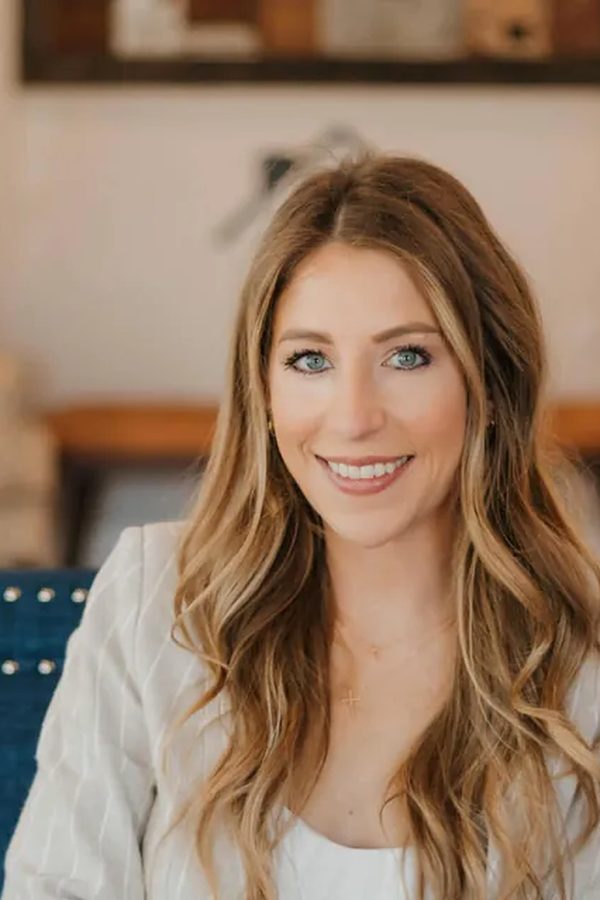 Brooke Pitkin, a smiling woman with long wavy blonde hair in a white pinstriped blazer, seated in a navy nailhead-trimmed chair.
