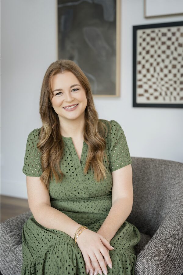 Sarah Hail, a smiling woman with long auburn hair in a green eyelet dress, seated in a grey armchair.